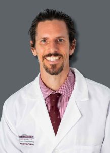 Man with white doctor coat and red tie smiling in front of a grey background