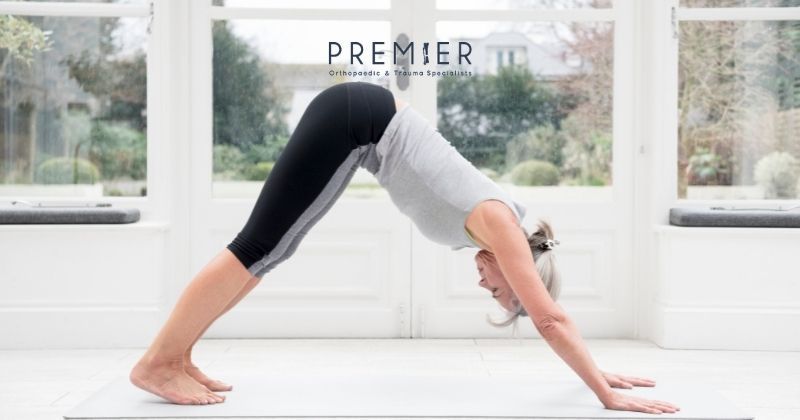 Silver haired woman wearing athletic attire in downward dog yoga position demonstrating exercises good for arthritis. Premier Orthopaedics logo overlay at top center. 