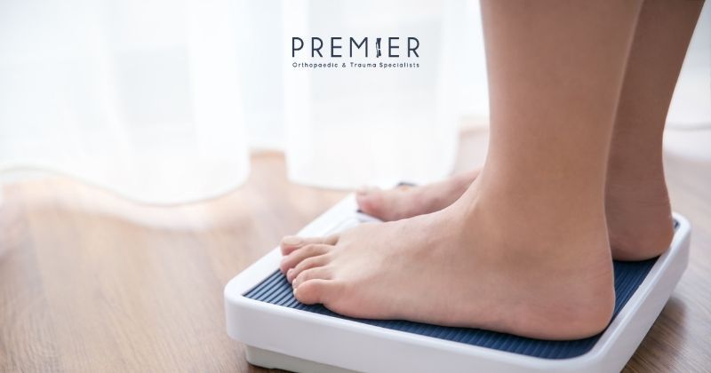 Premier Orthopaedics logo at top center of photo of feet on bathroom scale connecting pre-op weight loss and decreased surgical risk and improved orthopedic conditions.